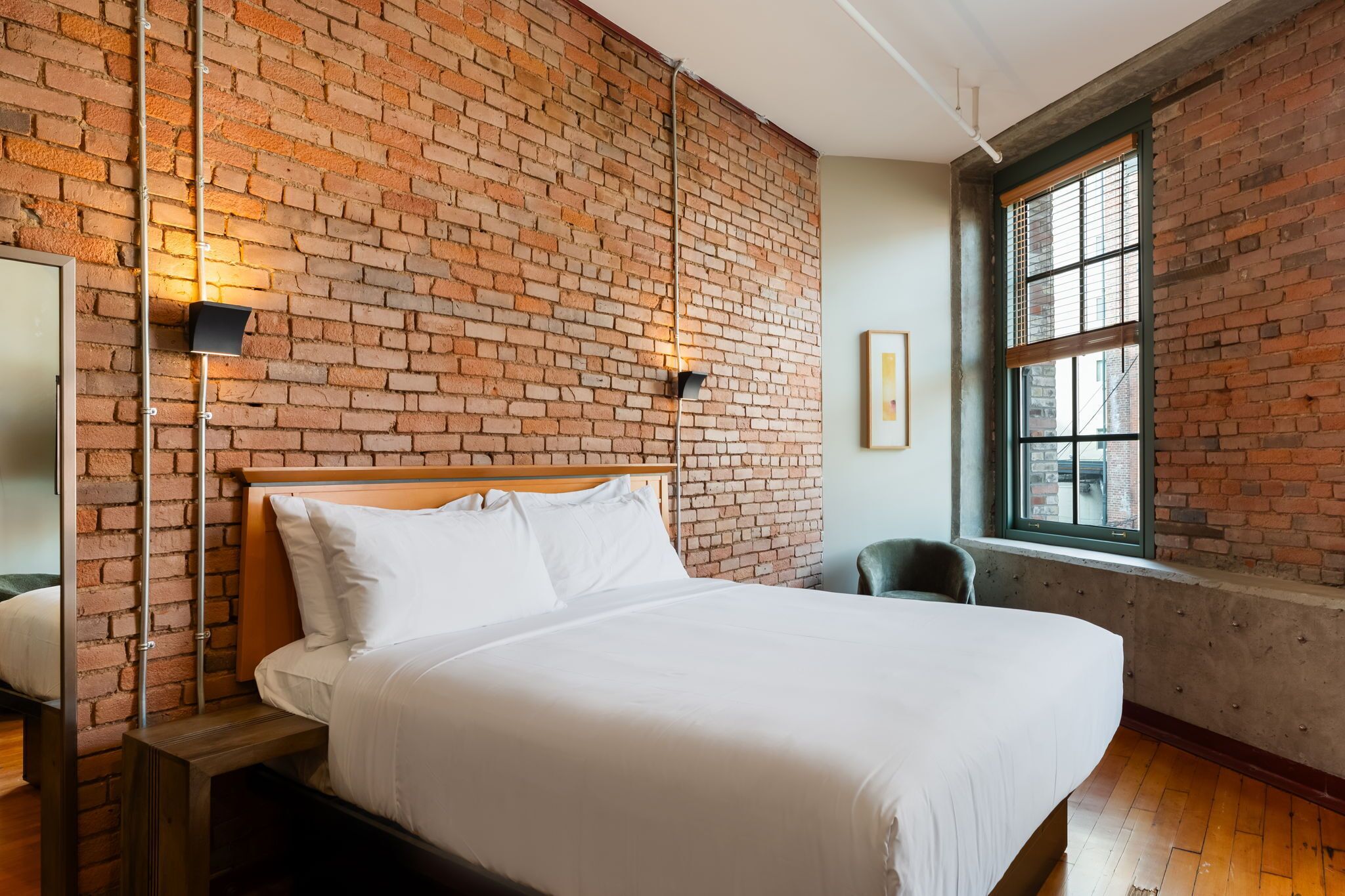 A hotel room with a brick wall, a large bed, a window with blinds, and a chair.