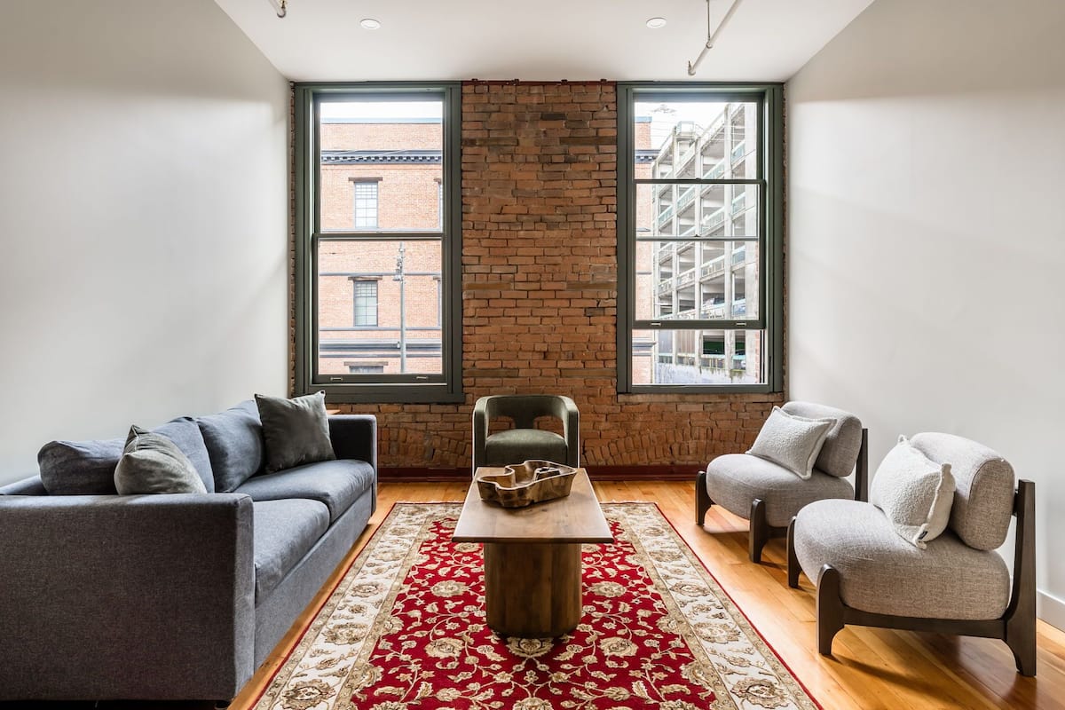 A living room with a brick wall, two windows, a sofa, a chair, and a coffee table.
