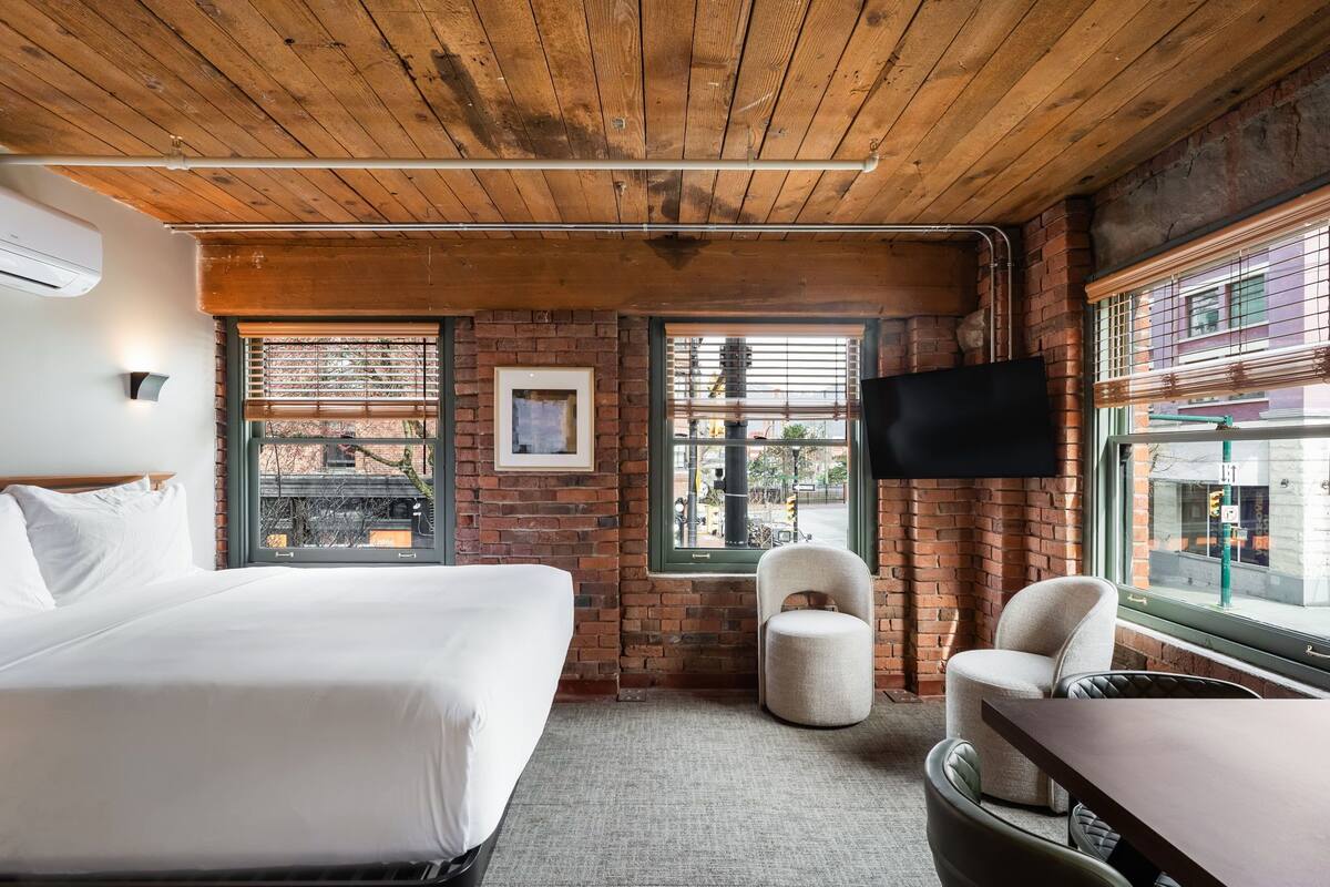 A hotel room with a wooden ceiling, brick walls, a bed, a chair, a table, and a TV mounted on the wall.