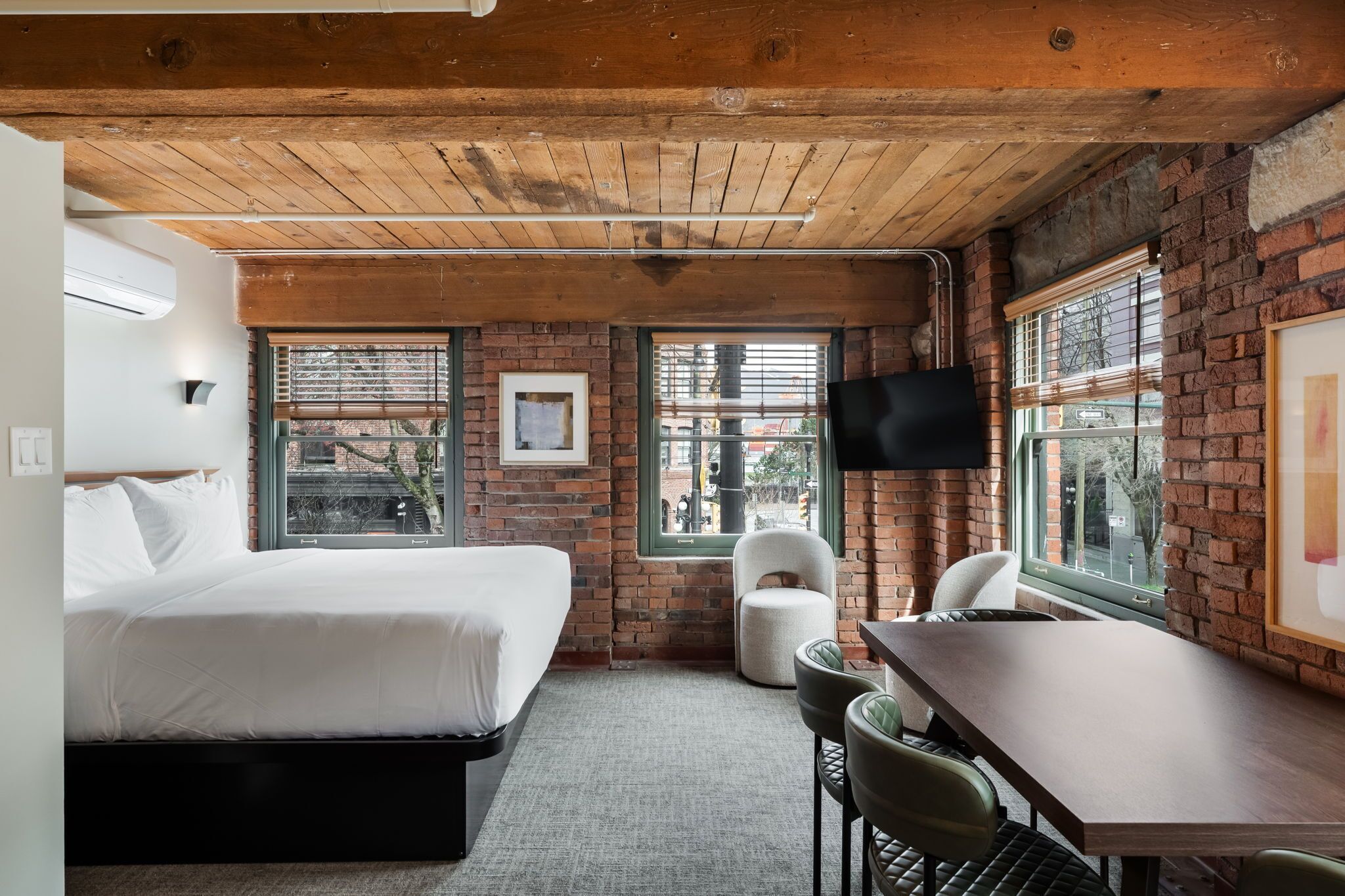A hotel room with a bed, a desk, two chairs, a TV, and large windows.