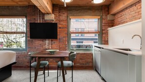 A compact kitchen with a brick wall, a flat-screen TV, a dining table with chairs, and a window with blinds. - Block House (Vancouver)