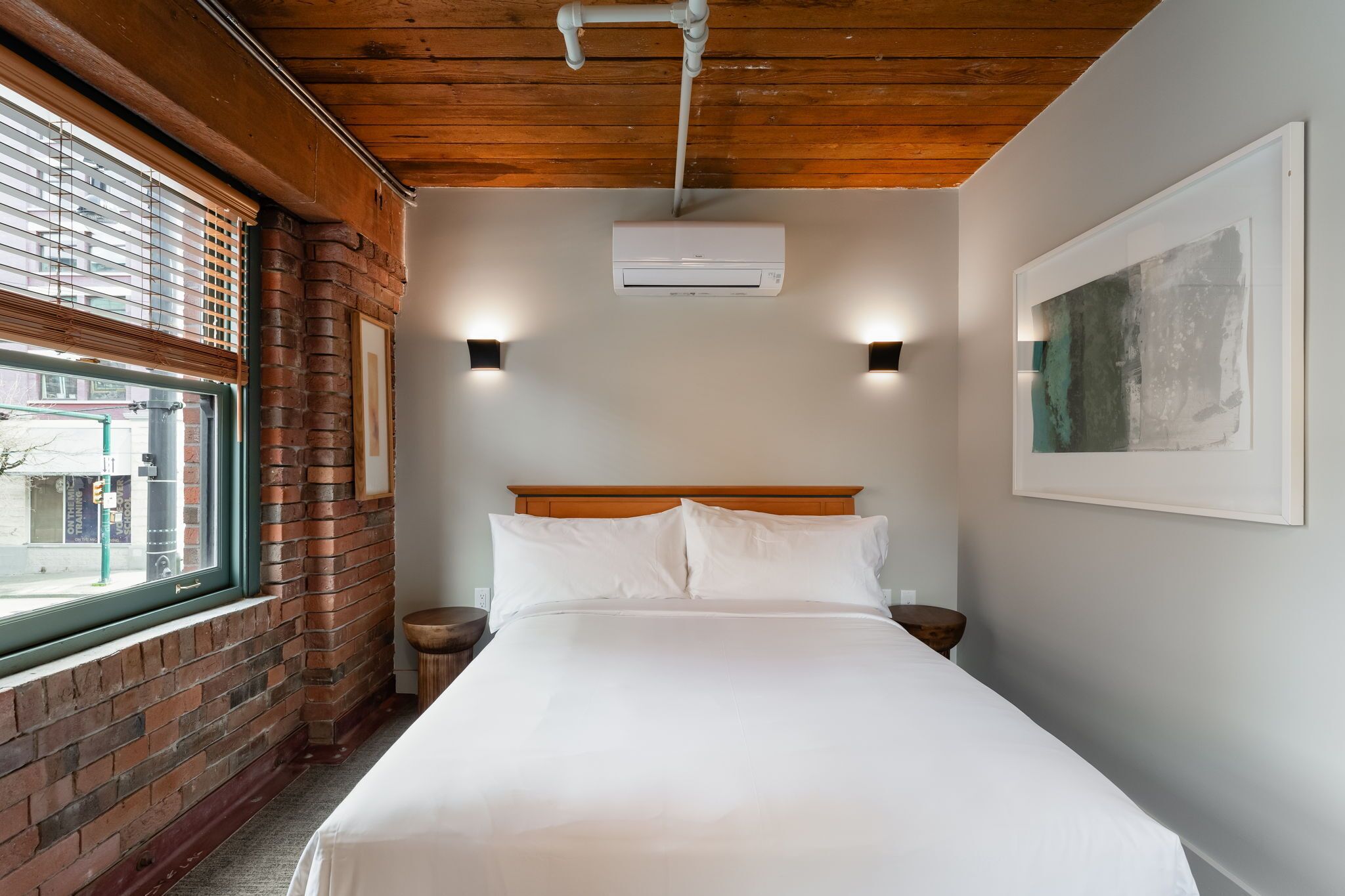 Ein Schlafzimmer mit Holzboden, einer Ziegelwand, einem Fenster, einem Bett mit weißer Bettwäsche, zwei Nachttischen und einer Klimaanlage.