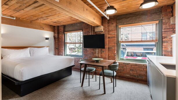 A bedroom with a bed, a desk, two chairs, a television, and a brick wall. - Block House (Vancouver)