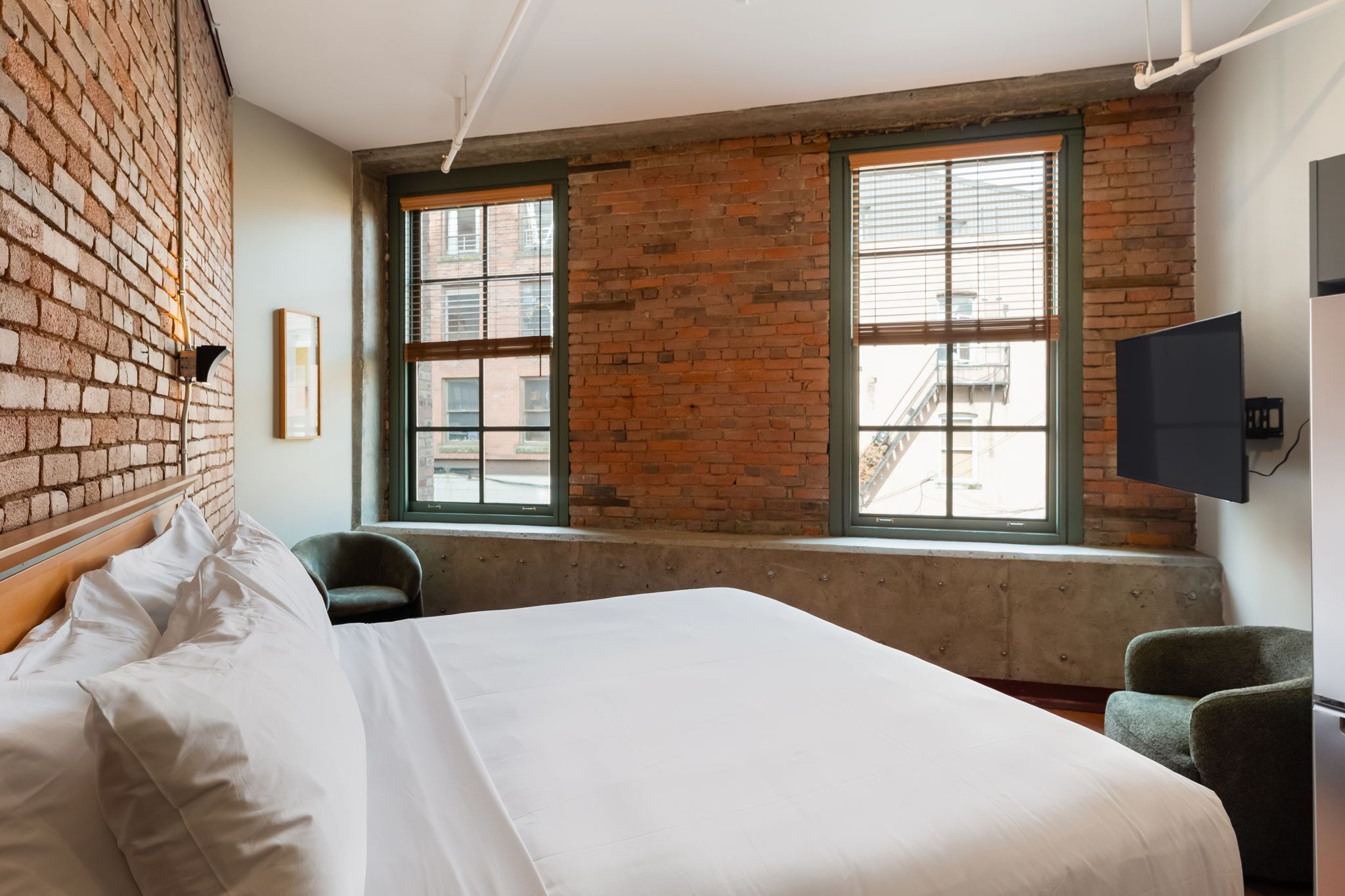 Een hotelkamer met een bakstenen muur, grote ramen, een bed, een stoel en een flatscreen televisie.