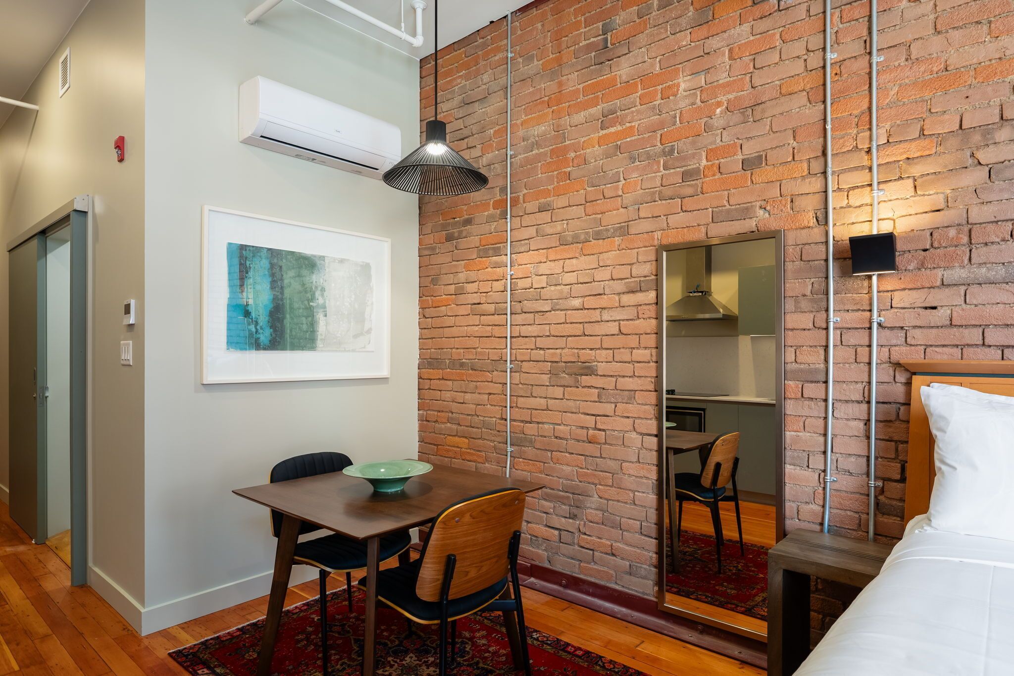A hotel room with a brick wall, a bed, a dining table with chairs, a small mirror, and a framed artwork.