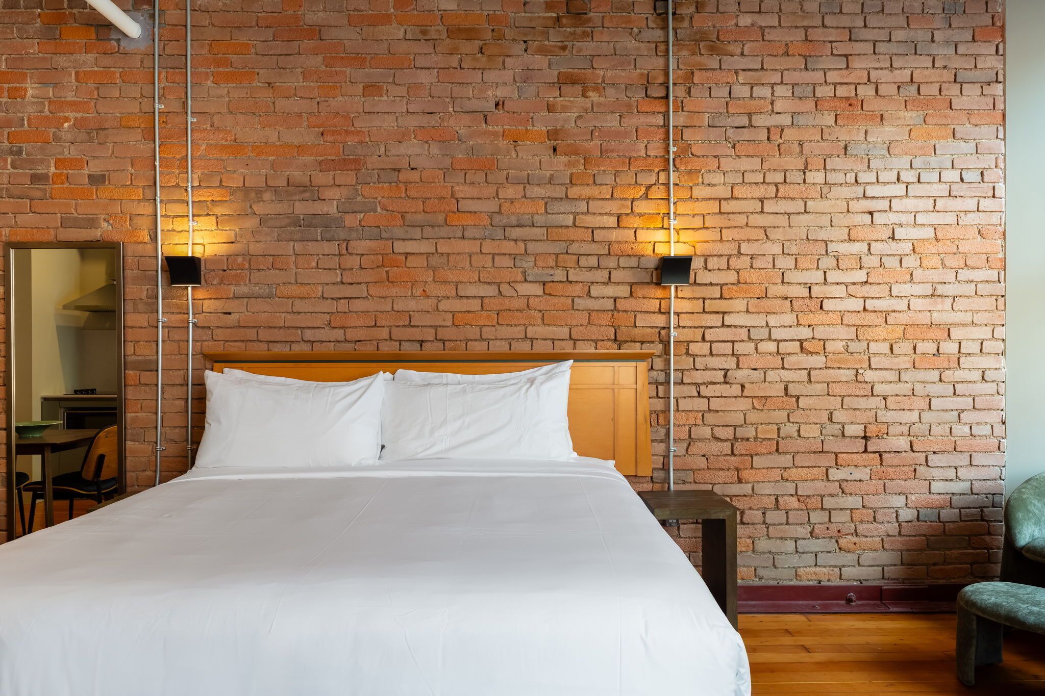 A hotel room with a brick wall, a wooden headboard, a bed with white linens, two wall-mounted lights, a small table, and a chair.