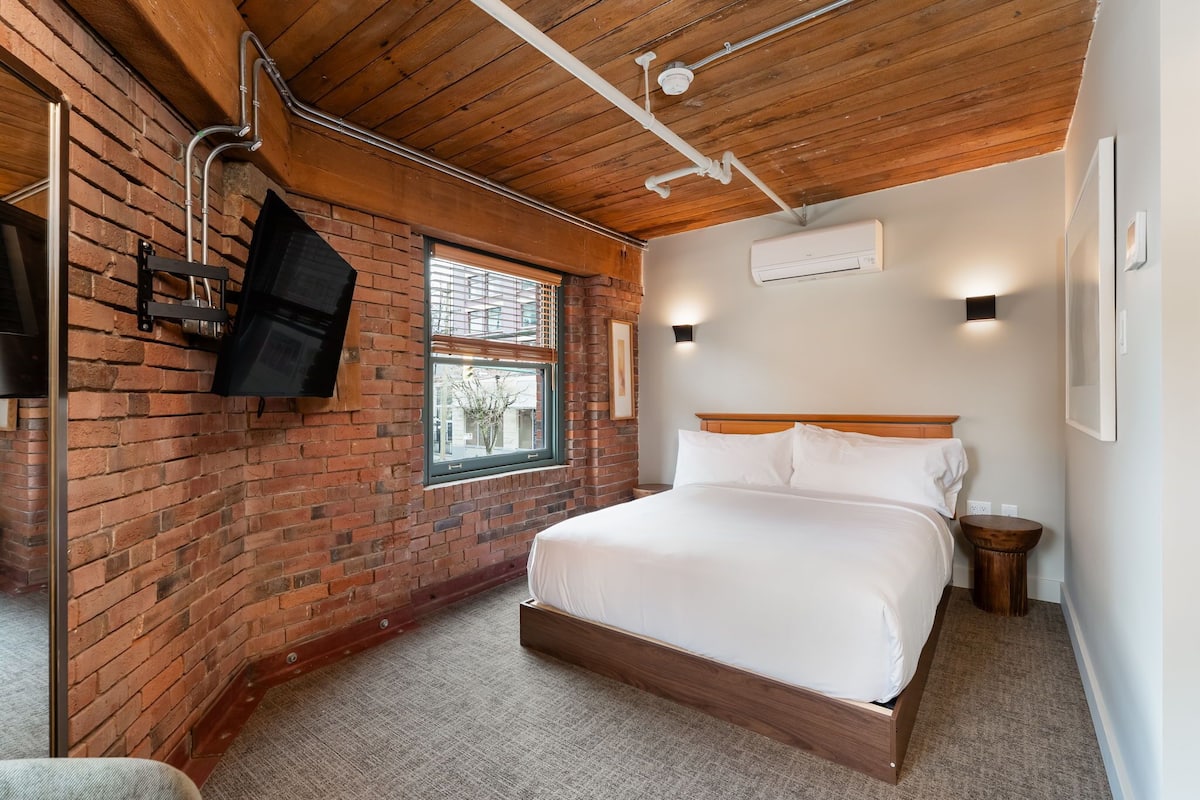 A bedroom with a brick wall, wooden ceiling, a bed, a TV mounted on the wall, and a window.