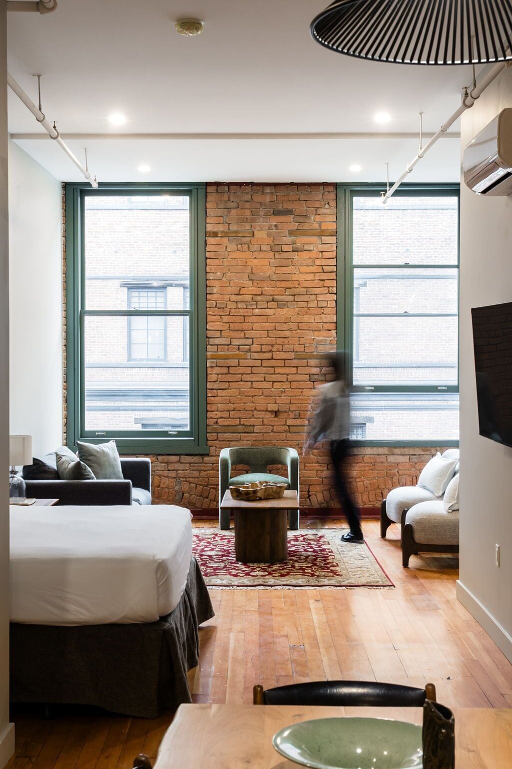 Une chambre moderne avec un mur en briques, de grandes fenêtres, un lit, une chaise, une table et un luminaire suspendu.