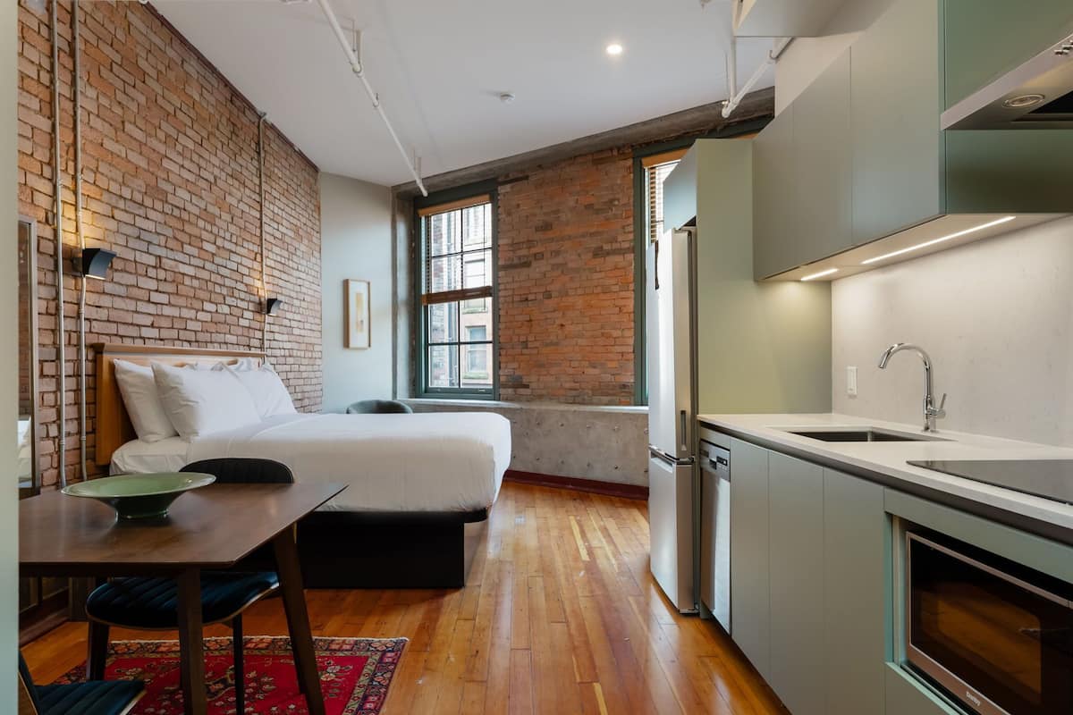 A compact room with a bed, a dining table, a kitchen area with a sink, and a brick wall.