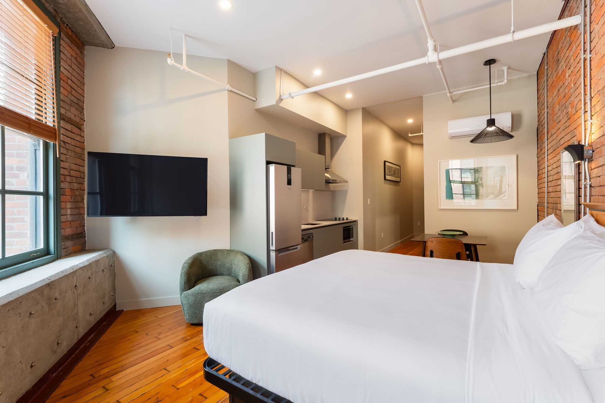 A modern hotel room with a large bed, a kitchenette, a seating area, and a TV mounted on the wall.