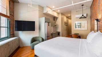 A modern hotel room with a large bed, a kitchenette, a seating area, and a TV mounted on the wall.