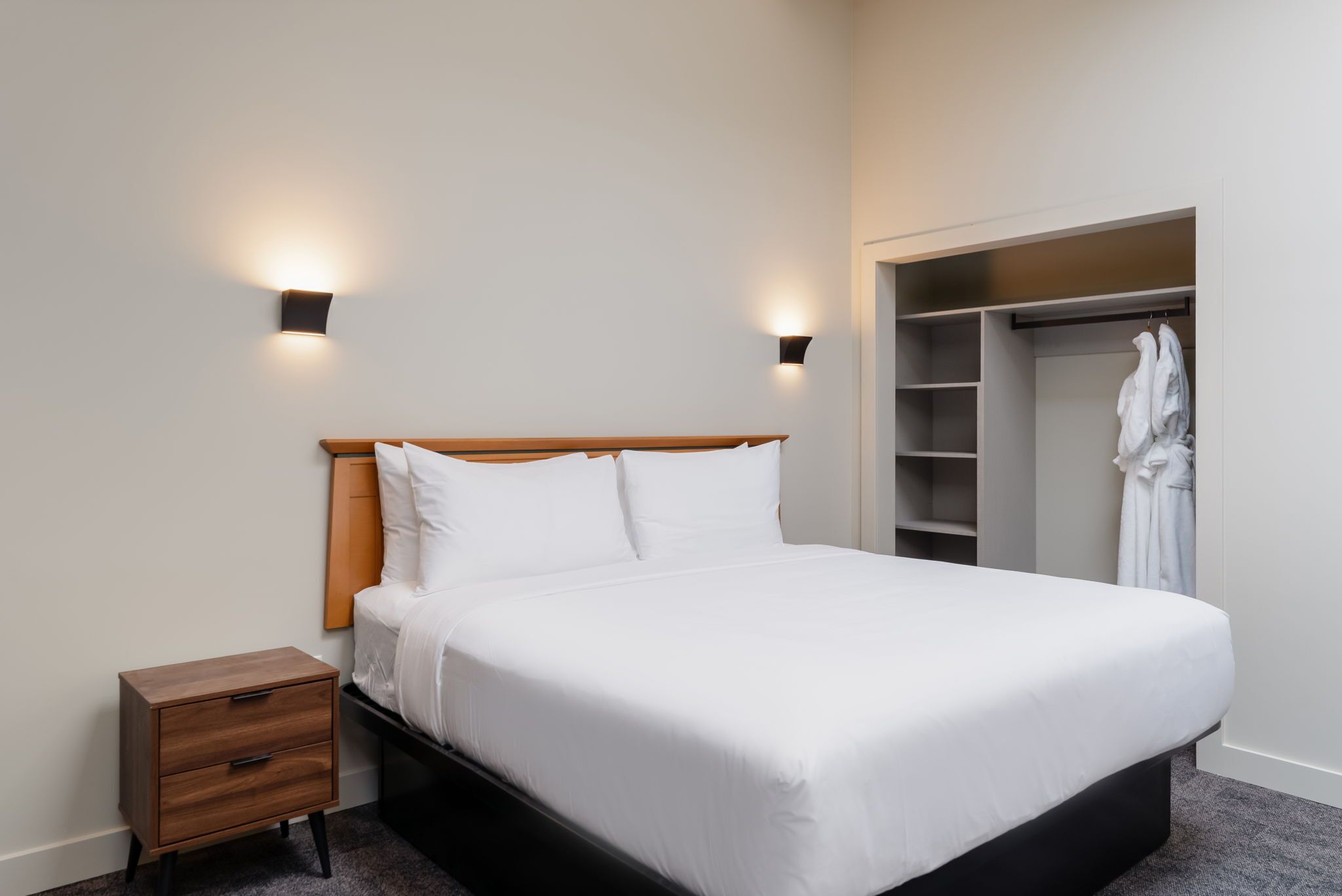 A neatly made bed with a wooden headboard, two bedside lamps, a small wooden nightstand, and a walk-in closet with hanging robes.