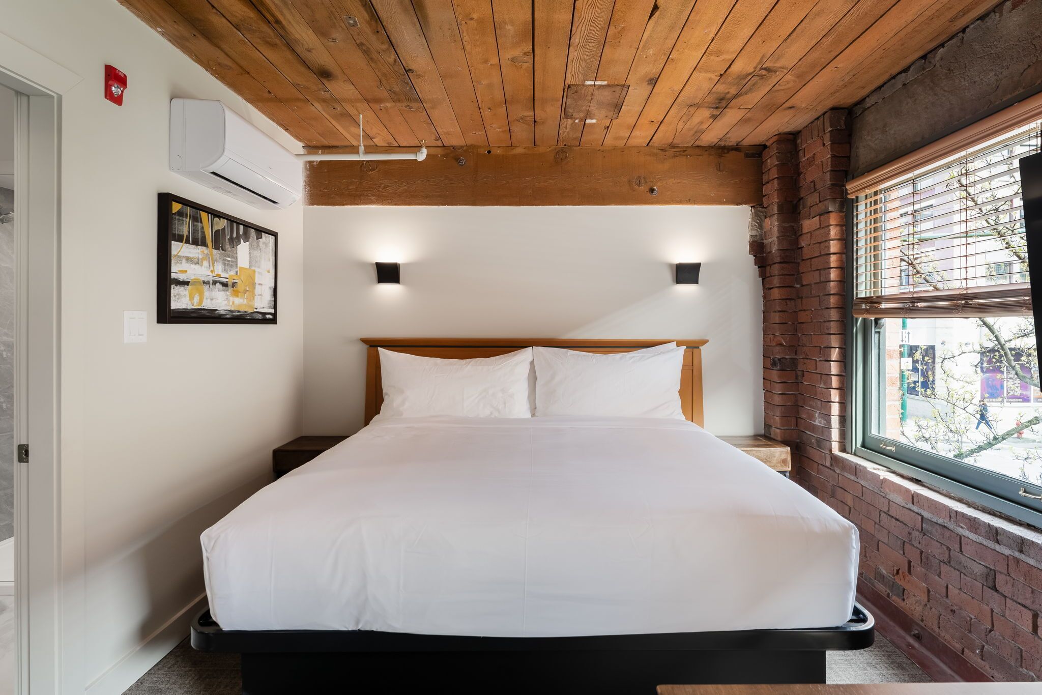 A bedroom with a wooden ceiling, a bed with white bedding, two wall-mounted lights, and a window with blinds.