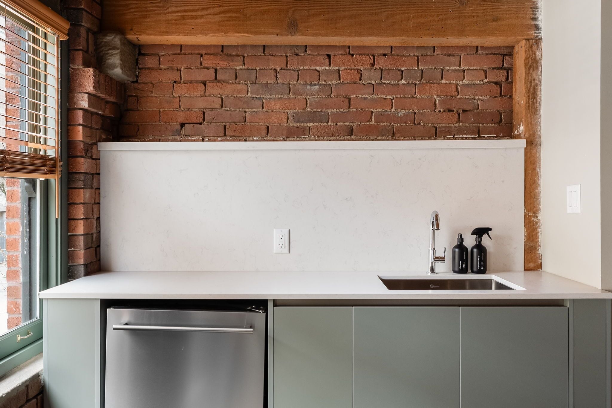 Una cocina con pared de ladrillo, encimera blanca, lavavajillas de acero inoxidable y un fregadero con dos botellas.