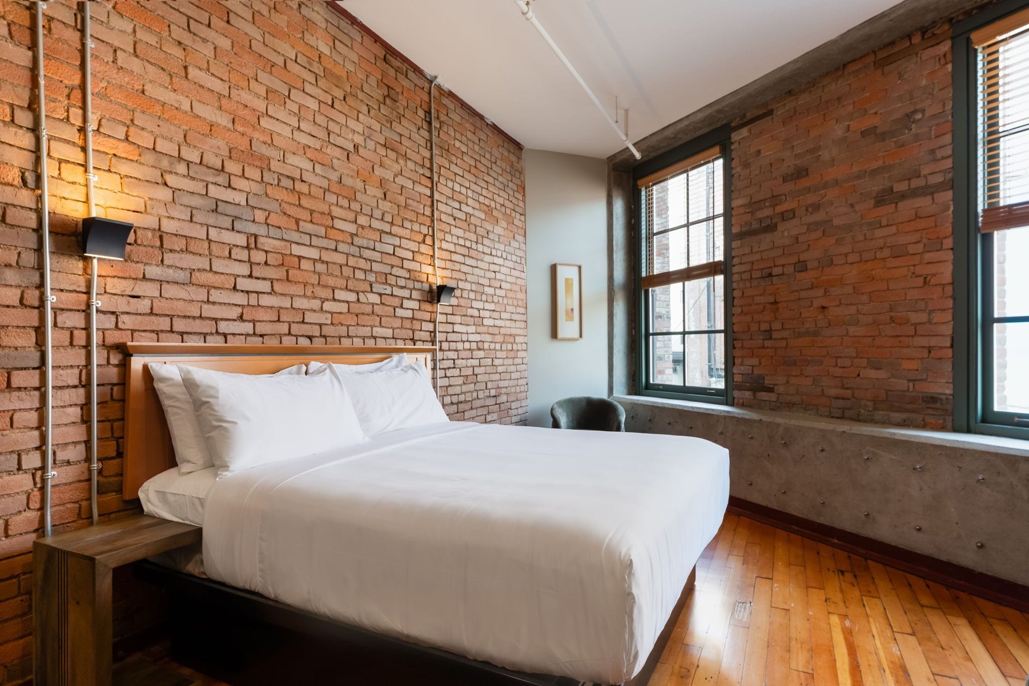 A hotel room with a brick wall, a bed, a chair, a window, and a picture on the wall.