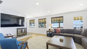 A spacious living room with a white brick fireplace, a flat-screen TV mounted above it, and a grey sofa. There are large windows with black frames, a wooden coffee table, and a blue armchair. - Golden Hour on Lake June - Dock, Deck & Views (Lake Placid)