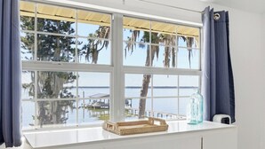 Une fenêtre donnant sur un quai et l’eau, des rideaux, un panier en osier et une bouteille en verre.