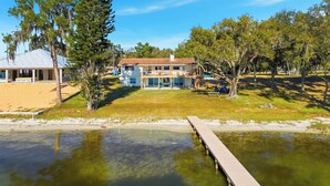 A two-story house with a dock extending into a calm body of water, surrounded by trees and a grassy area. - Golden Hour on Lake June - Dock, Deck & Views (Lake Placid)