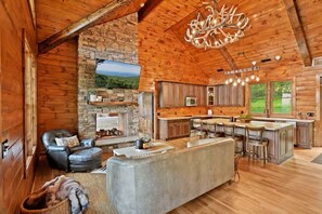 Un intérieur spacieux de chalet en bois avec une cheminée en pierre, une grande cuisine dotée de tabourets de bar et un coin salon confortable.