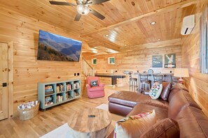 A cozy cabin interior with a wooden ceiling, a television mounted on the wall, a bookshelf, a leather sofa, and a dining area with a table and chairs. - New Cabin with Breathtaking View (Boone)