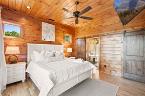 A bedroom with a large bed, a ceiling fan, wooden walls, a window, and a sliding door. - New Cabin with Breathtaking View (Boone)