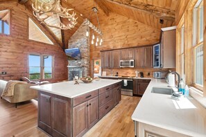 A spacious kitchen with a large island, wooden cabinetry, and a stone fireplace. - New Cabin with Breathtaking View (Boone)