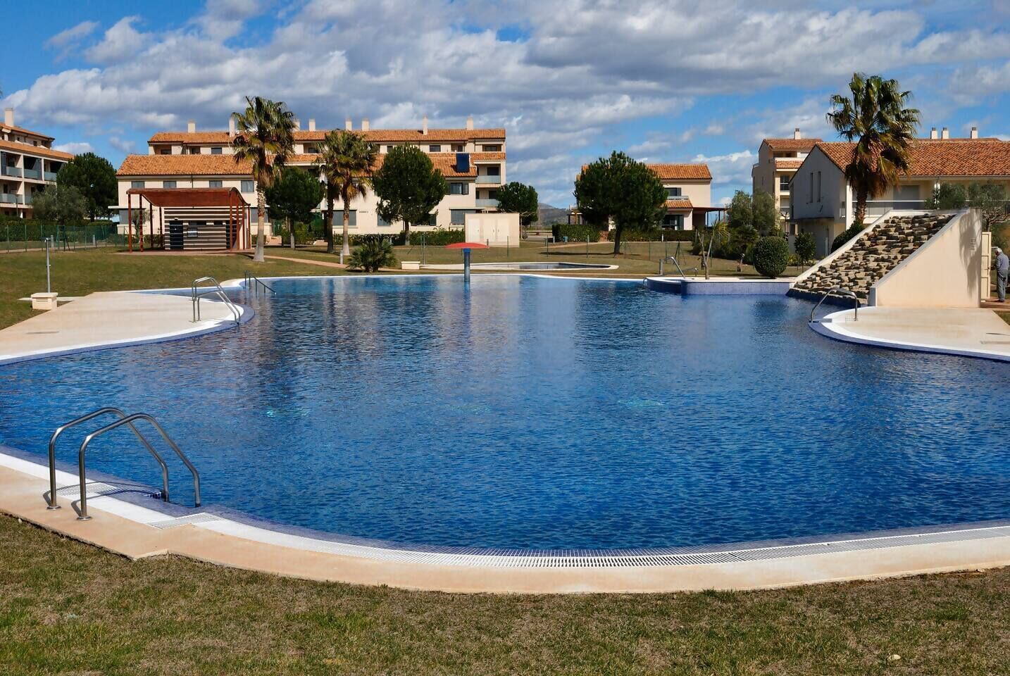 Una piscina con agua cristalina azul, rodeada por una solana de hormigón y adyacente a un edificio con tejado de azulejos.