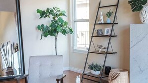 Uma sala de estar com uma poltrona cinza, uma mesa de centro redonda de madeira com livros, uma estante de metal preta com plantas e um espelho grande.