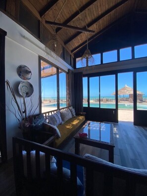 Ein geräumiger Raum mit großem Fenster zum Strand, Holzbalkendecke und einer Sitzecke mit Kissen.