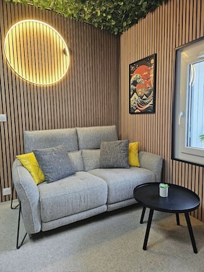 Un salon moderne avec un canapé gris, des coussins jaunes, une table ronde et un tableau de style japonais.