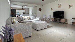 A modern living room with a grey sofa, ottoman, and a flat-screen TV on a wooden cabinet. There is a kitchen in the background with white cabinets and a hanging light fixture.