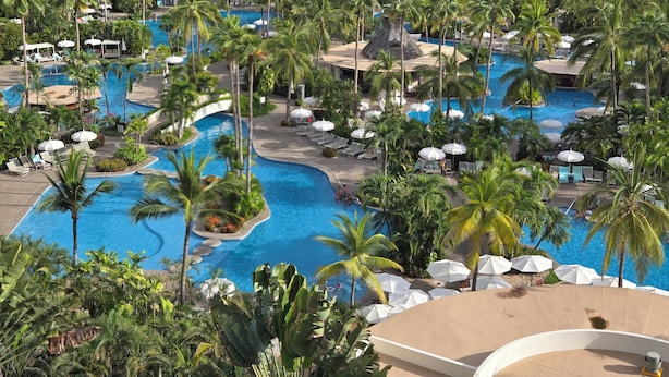 Ein Resort mit mehreren Schwimmbecken, umgeben von Palmen und Liegestühlen.