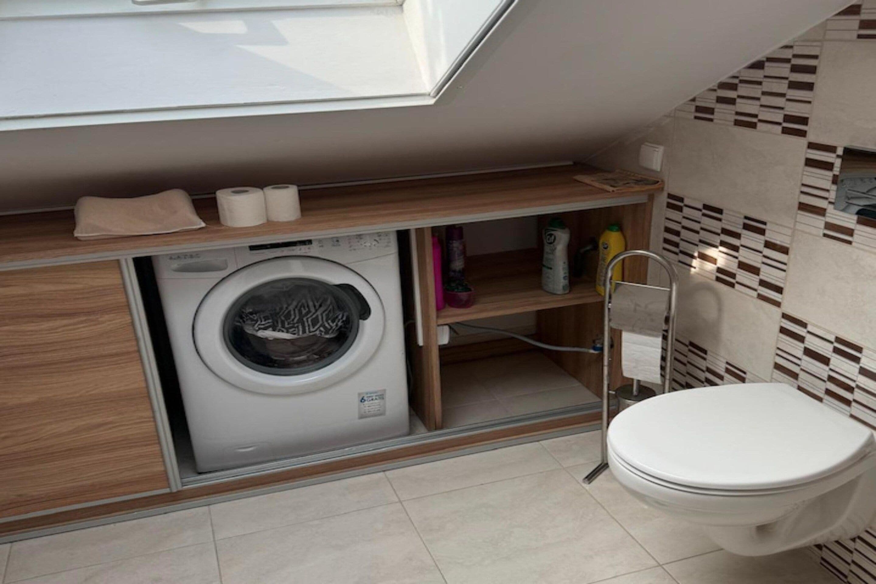Ein kleines Badezimmer mit Waschmaschine, Toilette und Ablage für Handtücher und Pflegeprodukte.