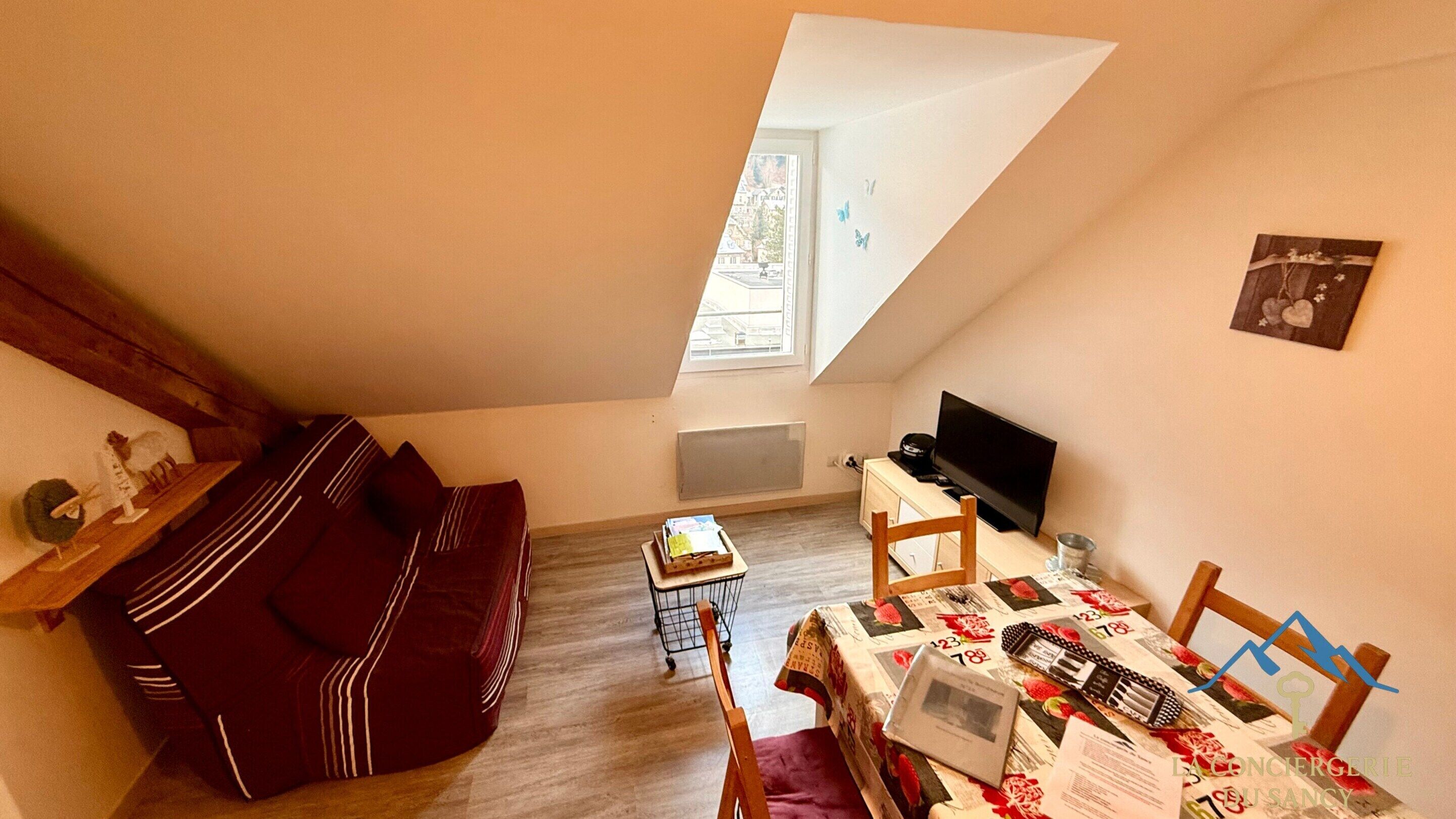 A small, cozy room with a sloped ceiling, a sofa, a table with a floral tablecloth, a television, and a window with a view of buildings.
