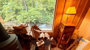 Ein gemütliches Blockhaus-Interieur mit Blick auf einen Wald und einen Bach, eine Holzkommode mit einer Lampe und ein Kamin mit Holzscheiten.