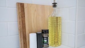 A wooden cutting board, a white cylindrical object, a black cylindrical object, a bottle of olive oil, and a spice container.