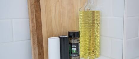 A wooden cutting board, a white cylindrical object, a black cylindrical object, a bottle of olive oil, and a spice container.
