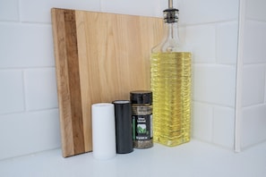 A wooden cutting board, a white cylindrical object, a black cylindrical object, a bottle of olive oil, and a spice container.
