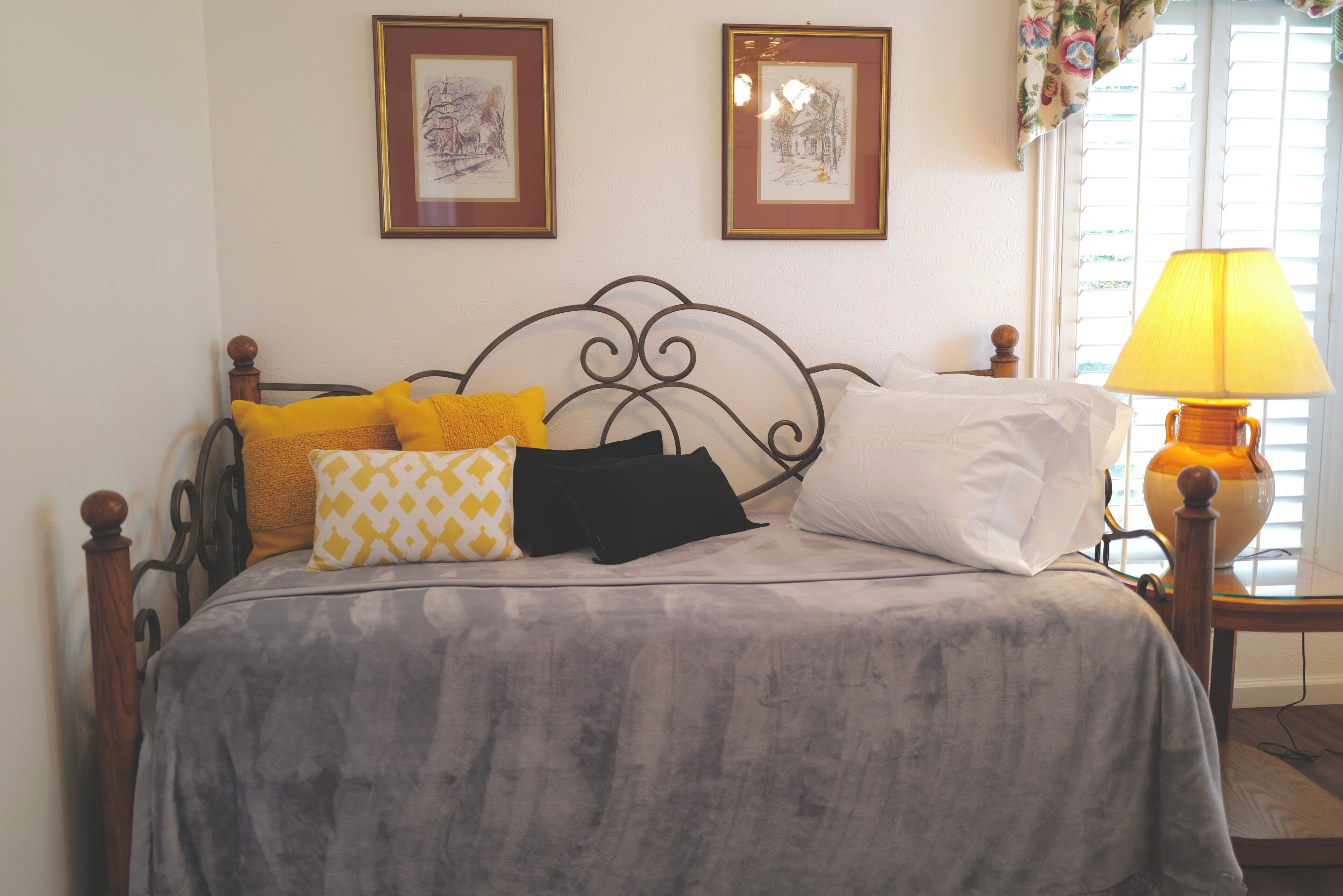 Cama com colcha cinzenta, várias almofadas e cabeceira de madeira. Duas obras de arte emolduradas estão penduradas na parede acima da cama.