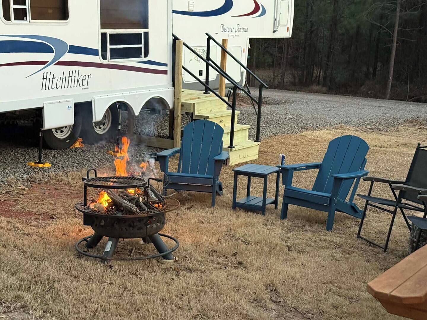 Un camping con un hoyo para asar, mobiliario exterior y una caravana que dice 'HitchHiker'.