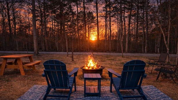 Ein Lagerfeuer mit zwei Adirondack-Stühlen, die darauf ausgerichtet sind, auf einem gemusterten Teppich in einem bewaldeten Gebiet bei Sonnenuntergang.