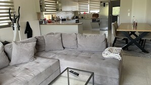 A modern living room with a grey sectional sofa, a glass coffee table, and a dining area with a wooden table and chairs. - 2-br Countryside Apartment in Mouzaki (Zakynthos)
