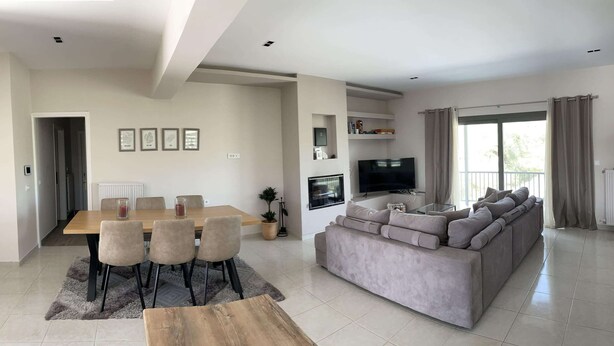 A modern living room with a large grey sectional sofa, a dining table with chairs, a flat-screen TV mounted on the wall, and a large window with curtains. - 2-br Countryside Apartment in Mouzaki (Zakynthos)