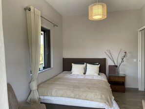 A bedroom with a bed, a bedside table, a hanging light fixture, a window with curtains, and a vase with branches. - 2-br Countryside Apartment in Mouzaki (Zakynthos)