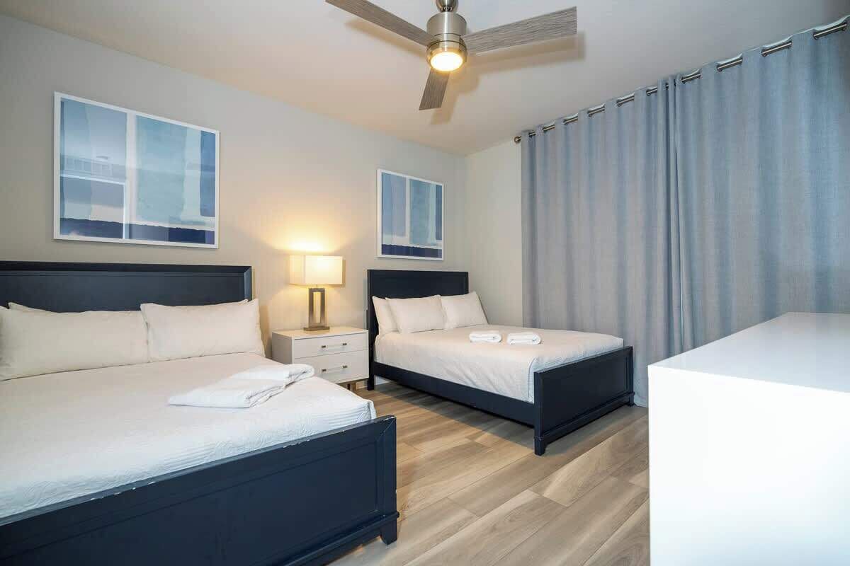 A hotel room with two beds, a ceiling fan, a nightstand, and a large window with curtains.
