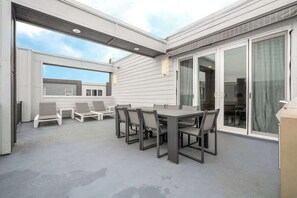 Un patio exterior con una mesa y sillas para comer, una zona de estar cubierta y un ventanal correderas hacia un espacio interior.