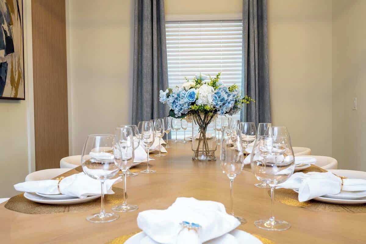Une table à manger dressée pour un repas, avec de nombreux verres à vin et des assiettes, un vase rempli de fleurs et, en arrière-plan, une fenêtre avec des stores.