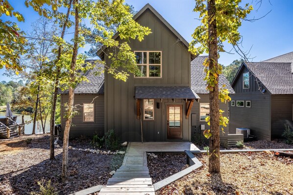 A two-story wooden house with a front porch, a small deck, and a landscaped yard with trees and a wooden walkway. - The Night Owl at Lake Martin - 3BR/3BA Home in Eclectic (Eclectic)