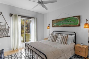 A bedroom with a bed, bedside table, lamp, and a window with curtains. - The Night Owl at Lake Martin - 3BR/3BA Home in Eclectic (Eclectic)