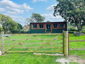 A green building with orange trim, a wooden gate, and a grassy area with trees and a table. - Cosy cabin, w/modern comforts minutes away from popular beaches & seaside towns. (Old Romney)